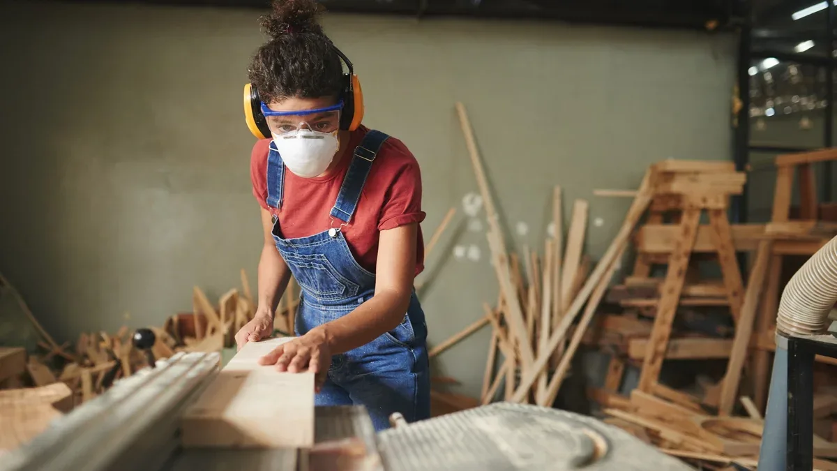Charpentière professionnelle en lunettes de protection, cache-oreilles et masque de découpe de planche de bois sur table de scie