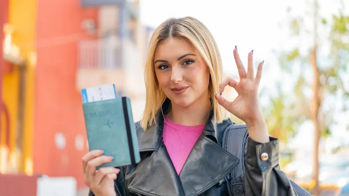Jeune jolie femme blonde tenant un passeport à l'extérieur montrant un signe ok avec les doigts